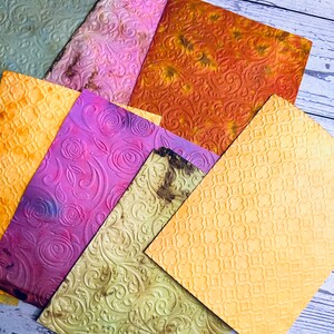 May include: A collection of textured, colorful paper sheets. The papers feature embossed patterns, including floral and geometric designs. Colors range from green, yellow, and orange to purple and pink, with a distressed appearance.