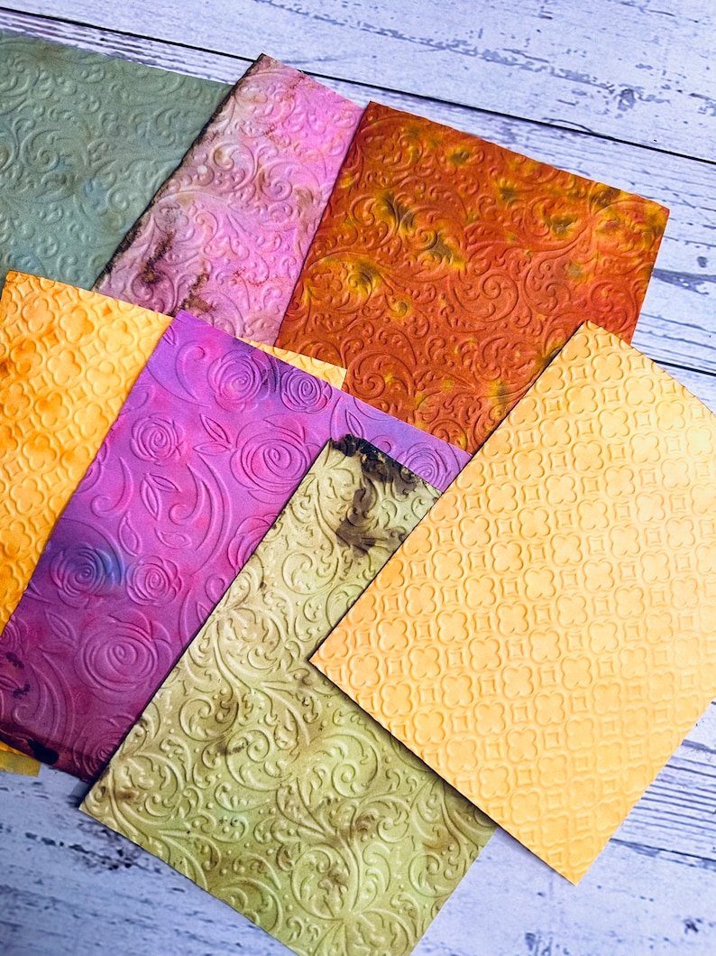 May include: A collection of textured, colorful paper sheets. The papers feature embossed patterns, including floral and geometric designs. Colors range from green, yellow, and orange to purple and pink, with a distressed appearance.