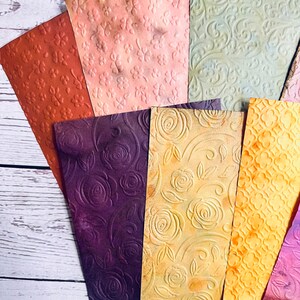May include: A collection of textured paper sheets in various colors, including orange, pink, green, and purple. Each sheet features an embossed floral or patterned design, creating a tactile and visually appealing effect. The papers are arranged on a white wooden surface.