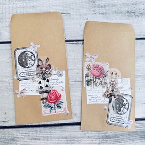 May include: Two brown paper envelopes decorated with vintage-style paper ephemera. One envelope features a black and white illustration of a girl in a dress with a crown, a red rose, and a moon. The other envelope features a black and white illustration of a girl in a dress with a crown, a rose, and a moon.