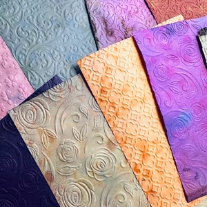 May include: A collection of textured paper sheets in various colors, including purple, orange, and green. Each sheet features embossed floral or geometric patterns. The papers have a vintage, aged appearance, with subtle color variations and distressed edges.