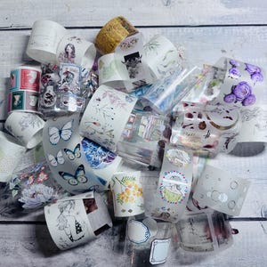 May include: A collection of decorative washi tape rolls with various designs, including butterflies, flowers, and illustrations. The tape features a range of colors, including blue, pink, and yellow, and is arranged on a light wood surface.