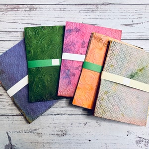 May include: A collection of colorful, textured paper packs. Each pack is wrapped with a band. The papers feature embossed floral and geometric patterns in shades of purple, green, pink, orange, and gray.