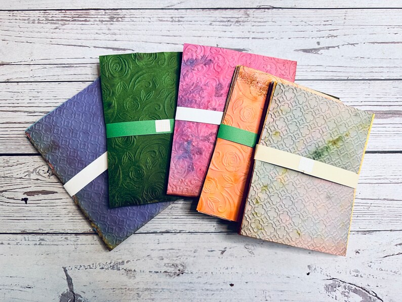 May include: A collection of colorful, textured paper packs. Each pack is wrapped with a band. The papers feature embossed floral and geometric patterns in shades of purple, green, pink, orange, and gray.