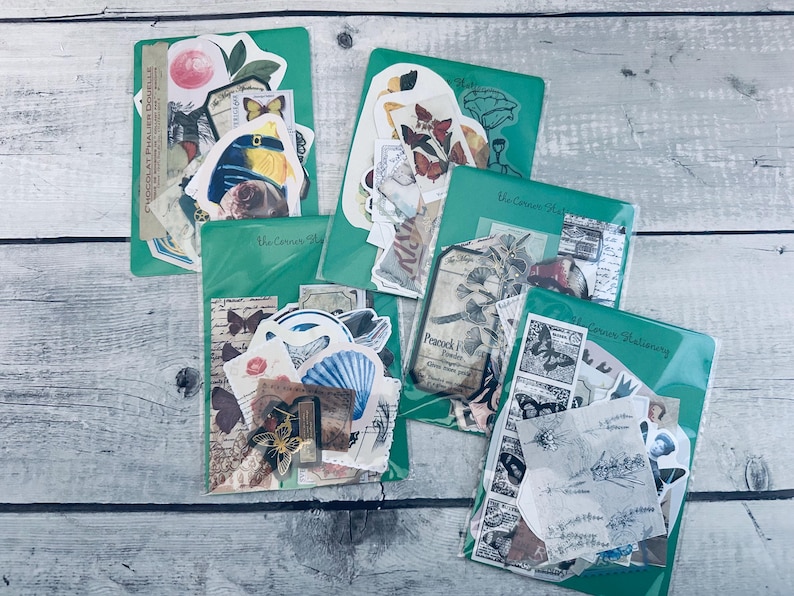 May include: Five clear plastic bags filled with various vintage style paper ephemera, including butterflies, flowers, and other decorative elements. The bags are labeled with the text "The Crown Shop" and "Crown Stationery".