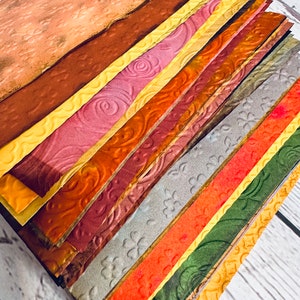 May include: A stack of textured paper sheets in various colors, including shades of brown, yellow, pink, orange, and green. The paper has embossed patterns of swirls, flowers, and other designs. The sheets are arranged diagonally, showcasing the different textures and colors.