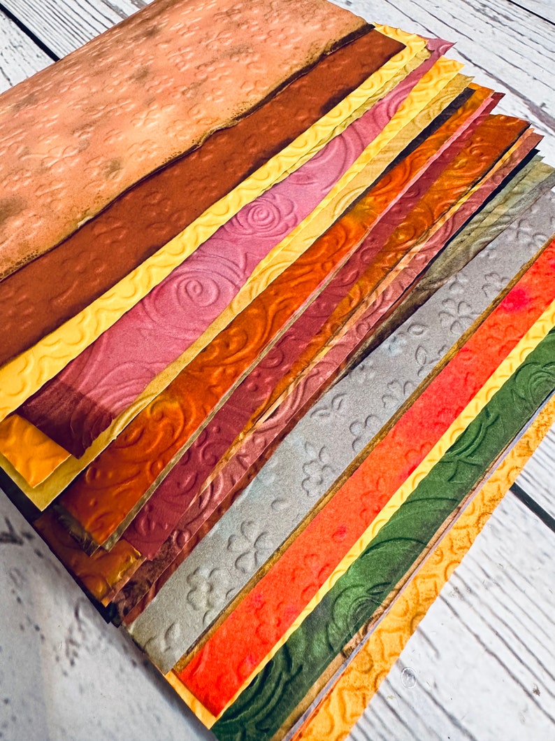 May include: A stack of textured paper sheets in various colors, including shades of brown, yellow, pink, orange, and green. The paper has embossed patterns of swirls, flowers, and other designs. The sheets are arranged diagonally, showcasing the different textures and colors.
