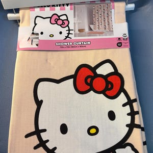 May include: A beige shower curtain featuring a large graphic of Hello Kitty with a red bow. The curtain is 72 inches by 72 inches. The packaging includes a smaller image of the curtain in a bathroom setting, and the words "Shower Curtain".