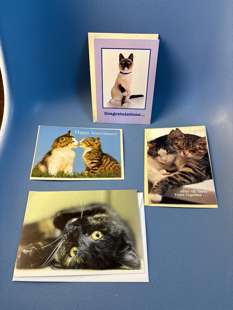 May include: Four greeting cards featuring cat images. One card says "Congratulations..." with a Siamese cat. Another says "Happy Anniversary!" with two cats kissing. A third card shows two cats cuddling, and the last card shows a black cat.