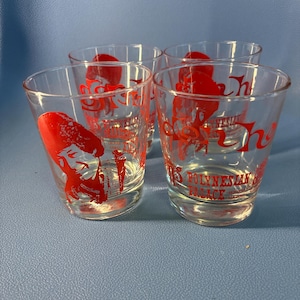 May include: A set of four clear glass shot glasses featuring red graphic prints. Each glass displays a stylized red illustration of a person and the red text Polynesian Palace Waikiki Hawaii printed on the front surface of the clear glass.