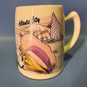 May include: Cream-colored ceramic mug featuring illustrations of seashells in shades of pink, yellow, and purple. The mug has a curved handle and the words "Atlantic City" are printed on the front.