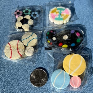May include: A collection of novelty erasers in various shapes, including soccer balls, baseballs, a paint palette, and planets. The erasers are packaged in clear plastic. A quarter is included for size comparison.