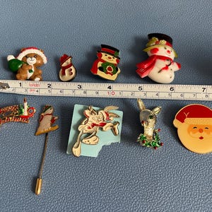 May include: A collection of Christmas-themed pins and brooches. Includes a bear with a sack, snowmen, a snowman, a Bugs Bunny pin, a rabbit with a wreath, and a Santa Claus face. A measuring tape is placed next to the pins.