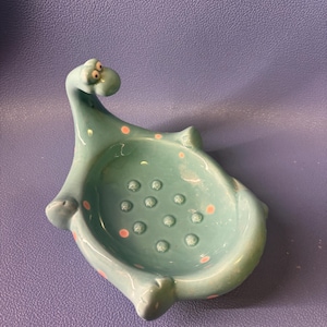 May include: A teal ceramic dinosaur-shaped soap dish with pink polka dots. The dish has a rounded bowl shape with small drainage holes and is set against a purple background. The dinosaur has a long neck and small arms.