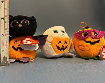 May include: Three Halloween-themed plush toys: a black bat with purple wings, a white ghost, and an orange pumpkin. The bat and pumpkin have jack-o'-lantern faces. Each toy has a tag. The bat measures approximately 3 inches.