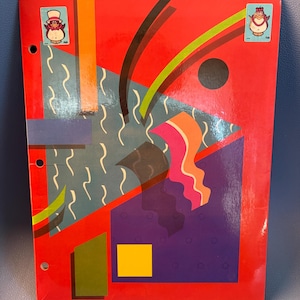 May include: A red binder with an abstract design featuring geometric shapes in orange, green, blue, and purple. Two stickers of a cartoon penguin wearing a top hat are on the cover. The binder has three holes for insertion into a ring binder.