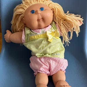 May include: A Cabbage Patch Kid doll with blonde hair, blue eyes, and a pink and green gingham dress. The doll is sitting on a blue chair.