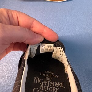 May include: Black slip-on shoe featuring a design from Tim Burton's "The Nightmare Before Christmas." The shoe has a white trim and a black and white pattern. The sole is brown.