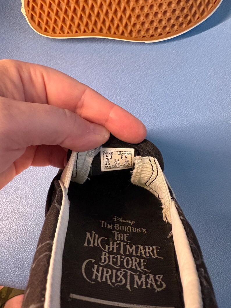 May include: Black slip-on shoe featuring a design from Tim Burton's "The Nightmare Before Christmas." The shoe has a white trim and a black and white pattern. The sole is brown.