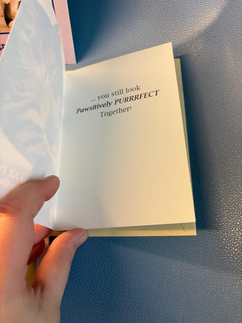 May include: A greeting card with the text "... you still look Pawsitively PURRRFECT Together!" The card is open, revealing the message on a cream-colored page. The background is a textured blue surface.