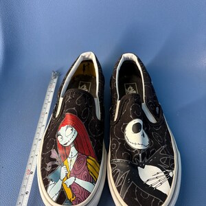 May include: Pair of black slip-on shoes featuring characters from The Nightmare Before Christmas. One shoe displays Sally, the other Jack Skellington, both with white soles and a black pattern.