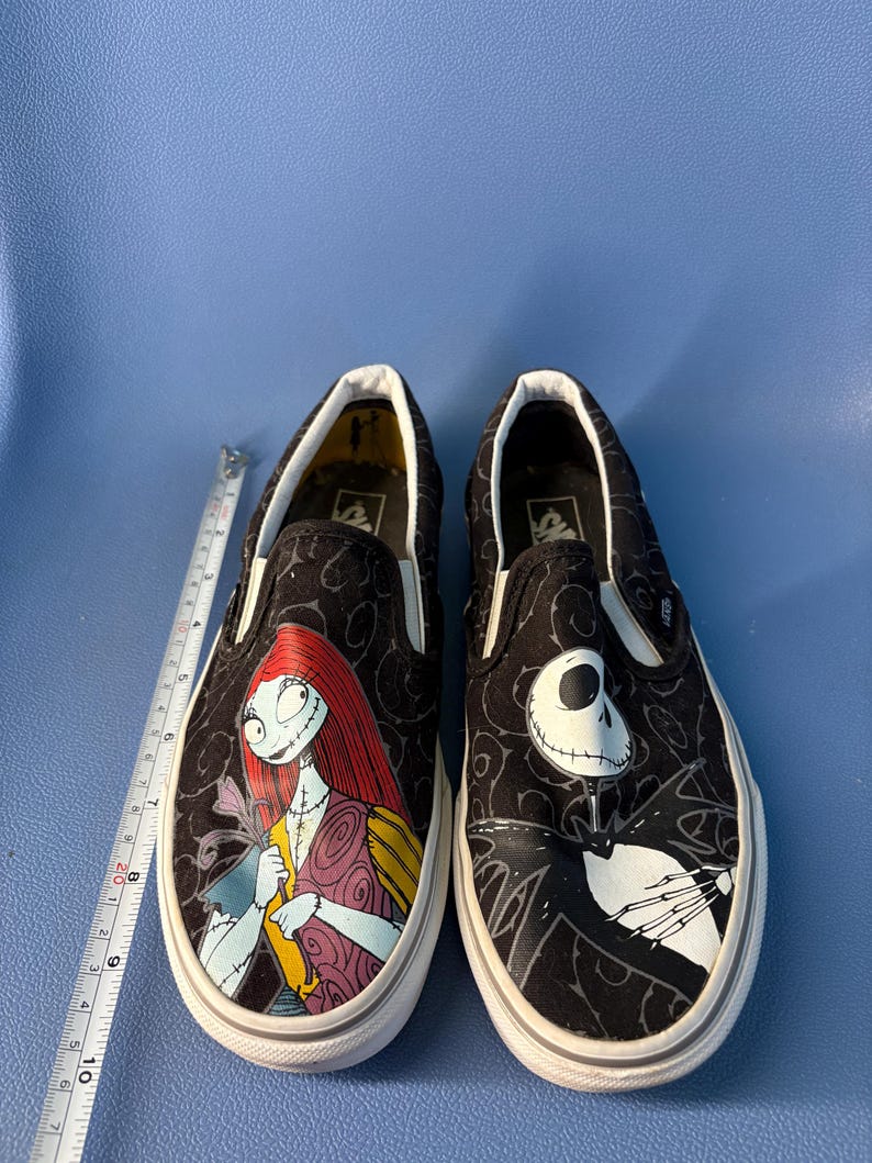 May include: Pair of black slip-on shoes featuring characters from The Nightmare Before Christmas. One shoe displays Sally, the other Jack Skellington, both with white soles and a black pattern.