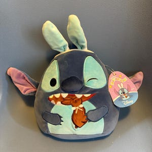 Puede incluir: Un personaje de Stitch de peluche y redondo, con orejas de conejo, detalles turquesa y un diseño de huevo de Pascua marrón en el vientre. El juguete tiene una etiqueta que dice "Squishmallows" y "Disney".