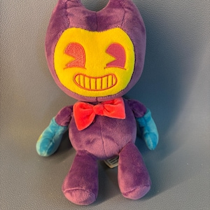 Può includere: Un peluche raffigurante un personaggio viola con un grande viso giallo, un ampio sorriso e occhi rosa. Il personaggio indossa un papillon rosso acceso al collo e ha le mani azzurre. Il peluche ha una consistenza morbida e un aspetto da cartone.