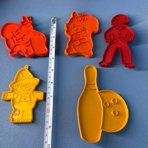 May include: Five colorful plastic cookie cutters in the shapes of a squirrel, a scarecrow, a bowling pin and ball, a person, and an elephant. The cookie cutters are orange and yellow. A ruler is next to the cookie cutters.