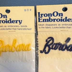 May include: Two packs of iron-on embroidery, each containing a white fabric patch with the word "Barbara" embroidered in yellow and blue thread. The packs are labelled "IronOn Embroidery" and "80¢".