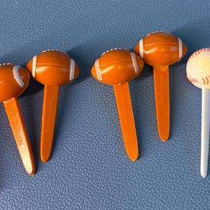 May include: Six sports-themed food picks including one orange basketball, four orange footballs with white laces, and one white baseball with red stitching. A white measuring tape is positioned on the right, showing a length of approximately 13.3 cm.