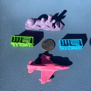 May include: A collection of colorful toy vehicles, including a purple train, a pink airplane, and two small train cars in green and blue. A coin is placed near the vehicles. The toys are made of a solid material.