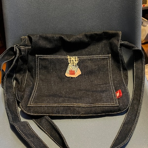 May include: A dark blue denim crossbody bag with a small patchwork doll applique on the front flap. The bag has a long adjustable strap and a small pocket on the front.