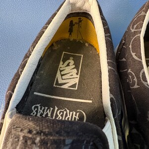 May include: Close-up of a pair of black slip-on shoes with a repeating pattern. The interior shows the Vans logo and the word "Christmas". The shoe's interior has a yellow accent.
