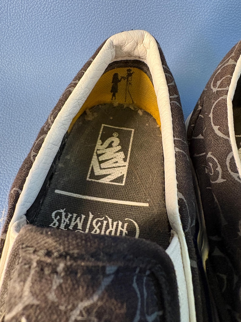 May include: Close-up of a pair of black slip-on shoes with a repeating pattern. The interior shows the Vans logo and the word "Christmas". The shoe's interior has a yellow accent.