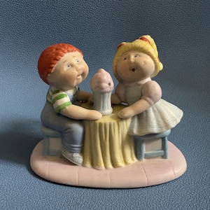May include: A ceramic figurine of two children at a table with a cupcake. The boy has red hair, a green and white striped shirt, and blue overalls. The girl has blonde hair and a light blue dress. The table has a yellow tablecloth.