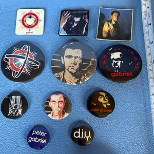 May include: A collection of Peter Gabriel and related band buttons and pins. The pins feature images of Peter Gabriel, album art, and the text "gabriel" and "d.i.y." The pins are various sizes and shapes, with a blue background.