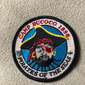 May include: A round, embroidered patch with a black border. The center features a cartoon pirate with a red eyepatch, black beard, and a yellow and black hat. The text "Camp Bicoco 1998 Pirates of the Seas" surrounds the pirate.