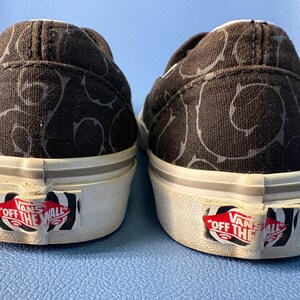 May include: Pair of black slip-on sneakers with a repeating gray swirl pattern. The shoes have white soles with a red and white "Off The Wall" logo on the heel.