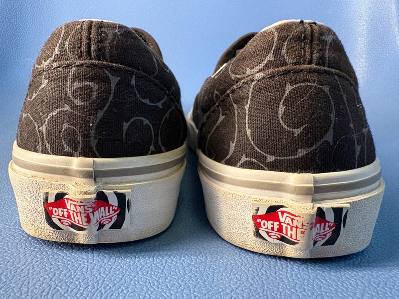 May include: Pair of black slip-on sneakers with a repeating gray swirl pattern. The shoes have white soles with a red and white "Off The Wall" logo on the heel.