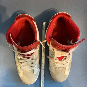 May include: A pair of white and red high-top sneakers with white laces. The sneakers are on a blue surface with a ruler for scale.