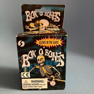May include: A box of glow-in-the-dark skeleton toys called "Box O Bones". The box features a cartoon skeleton with a skull and crossbones design. The box has a warning label that says "CHOKING HAZARD - Small parts. Not for children under 3 years."
