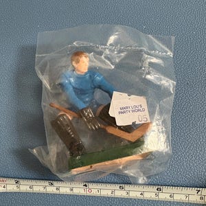 Könnte beinhalten: Eine Plastikfigur eines Hockeyspielers, der ein blaues Trikot und braune Hosen trägt und einen Hockeyschläger hält. Die Figur ist in einer durchsichtigen Plastiktüte verpackt, auf der ein Etikett mit der Aufschrift "Mary Lou's Party World" und "05" angebracht ist.