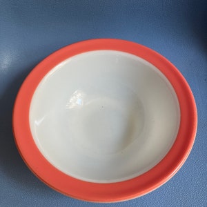 Può includere: Una ciotola in ceramica bianca con un bordo color corallo. La ciotola ha un design semplice e pulito ed è adatta per servire cibo. La ciotola è posta su uno sfondo blu.