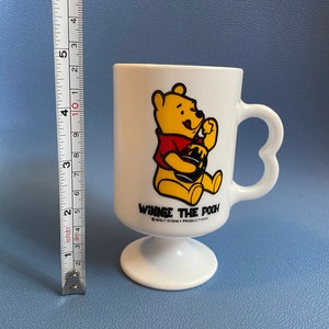 May include: White Winnie the Pooh pedestal mug. The mug features a yellow bear wearing a red shirt and holding a honey pot. The handle is heart-shaped. The mug is approximately 4 inches tall.