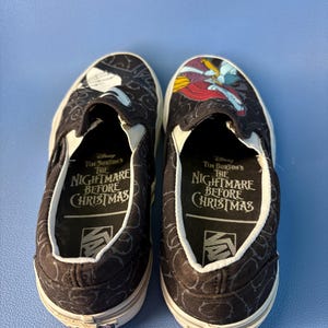 May include: Pair of black slip-on shoes featuring characters from Tim Burton's The Nightmare Before Christmas. The shoes have a white sole and a black and white pattern. The inner sole has the text "The Nightmare Before Christmas".