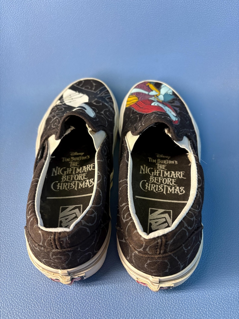 May include: Pair of black slip-on shoes featuring characters from Tim Burton's The Nightmare Before Christmas. The shoes have a white sole and a black and white pattern. The inner sole has the text "The Nightmare Before Christmas".