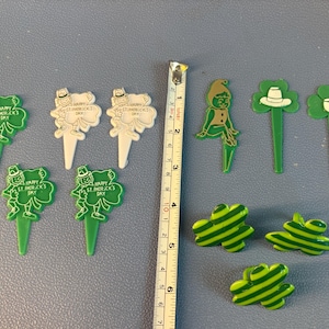 May include: A collection of green and white plastic St. Patrick's Day themed cupcake toppers and decorations, including leprechaun, shamrock, hat, and pipe shapes, displayed next to a yellow measuring tape showing approximately 5 inches in length.