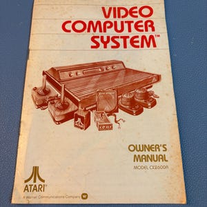 Puede incluir: Manual del propietario de la consola de videojuegos Atari CX2600. La portada presenta las palabras "VIDEO COMPUTER SYSTEM" en rojo, junto con una ilustración de la consola y los mandos. El logotipo de Atari es visible.