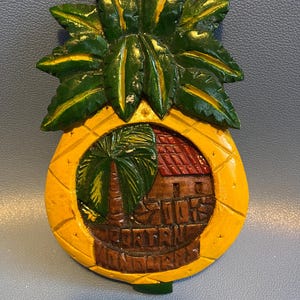 May include: A wooden pineapple-shaped decorative item. The top features green leaves with yellow accents. The center has a carved scene of a house and palm tree, with the text "700's PORT ANTONIO, JAMAICA".